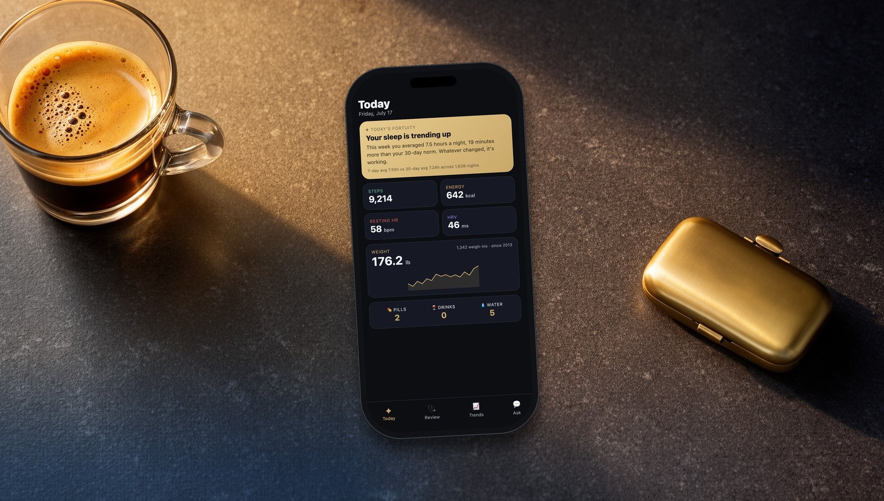 A smartphone showing the Fortuity Today screen, on a stone counter beside an espresso and a brass pill case
